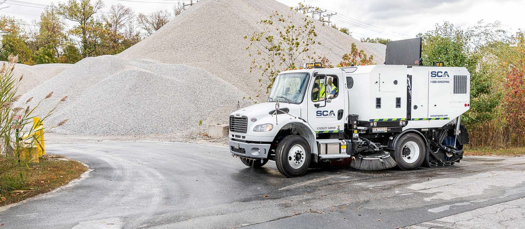 A sweeping truck cleaning the road near a large pile of gravel